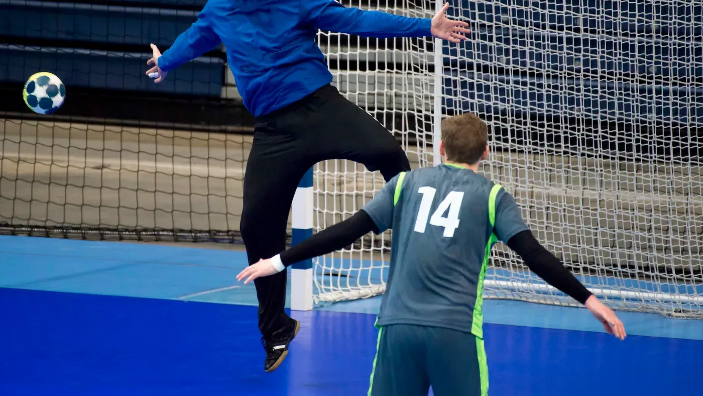 Handbollsmålvakt hoppar upp för att rädda ett skott medan en försvarare står framför målet