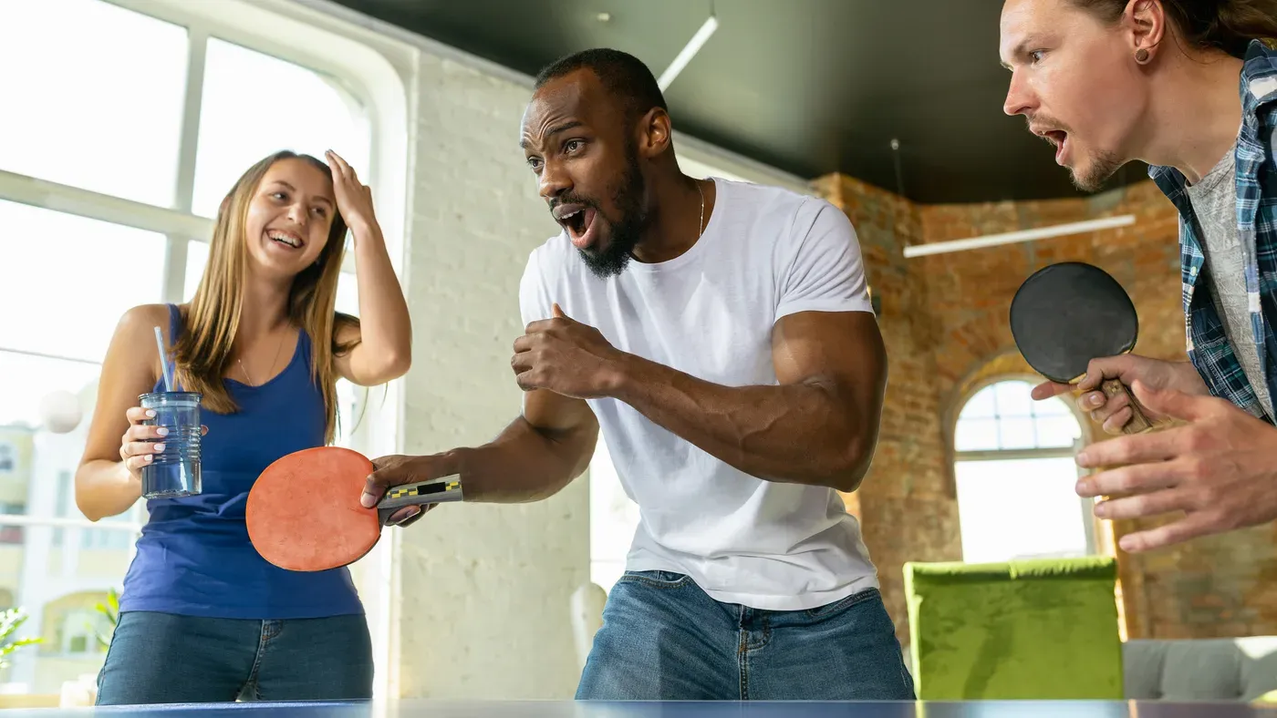 Glada vänner med pingisracketar spelar pingis tillsammans inomhus