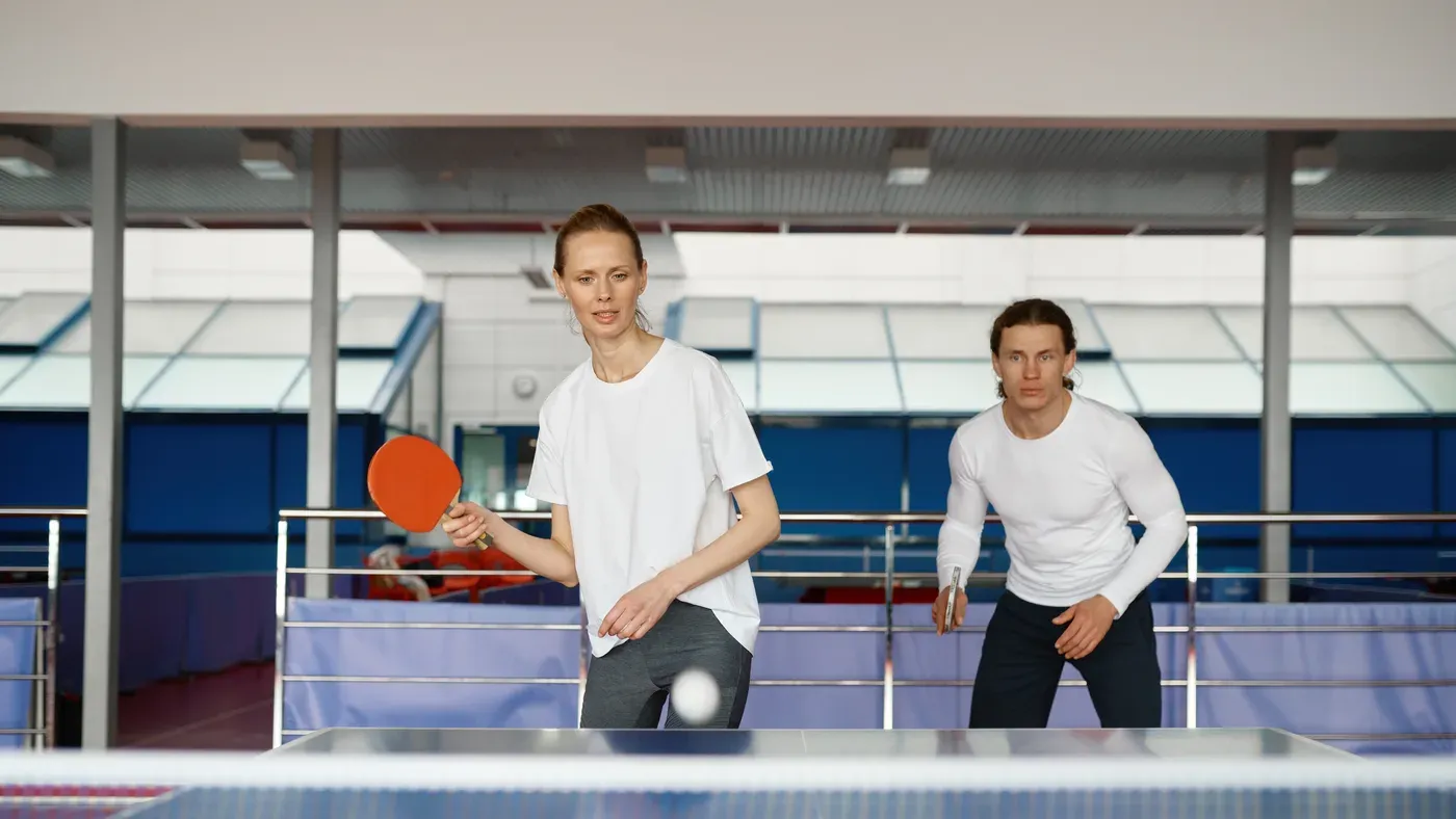 Två spelare spelar dubbel vid ett blått bordtennisbord i en hall